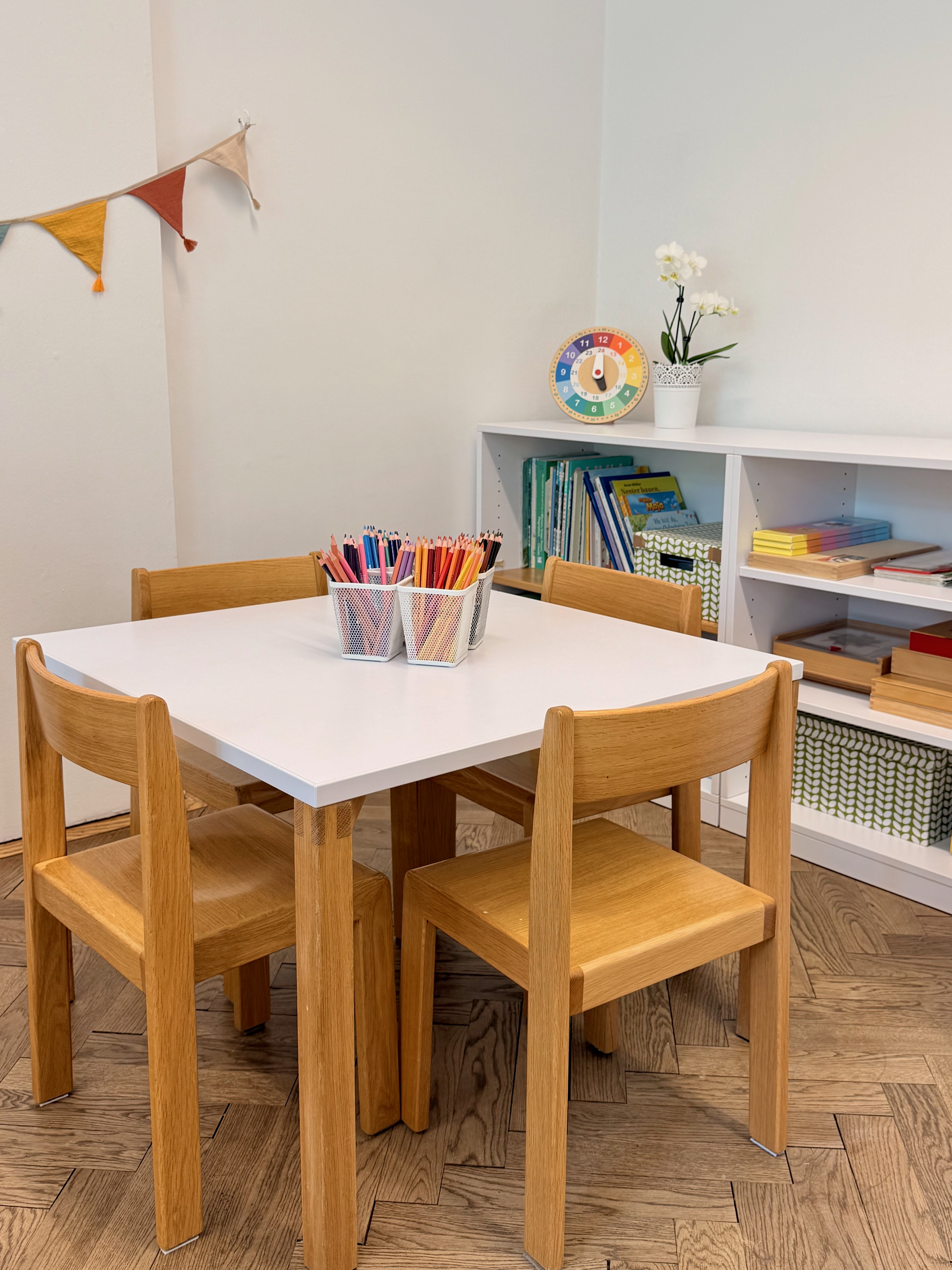 Kindertisch mit vier Holzstühlen, darauf Becher mit bunten Buntstiften. Im Hintergrund ein weißes Regal mit Büchern und Kisten, eine bunte Uhr und eine Blume, an der Wand eine Wimpelgirlande.