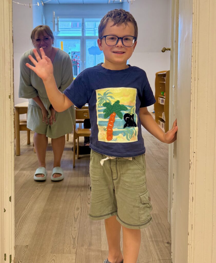Ein lächelnder Junge mit Brille steht im Türrahmen, im Hintergrund winkt eine Betreuerin.
