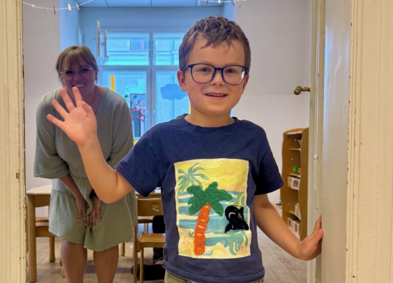 Ein lächelnder Junge mit Brille steht im Türrahmen, im Hintergrund winkt eine Betreuerin.