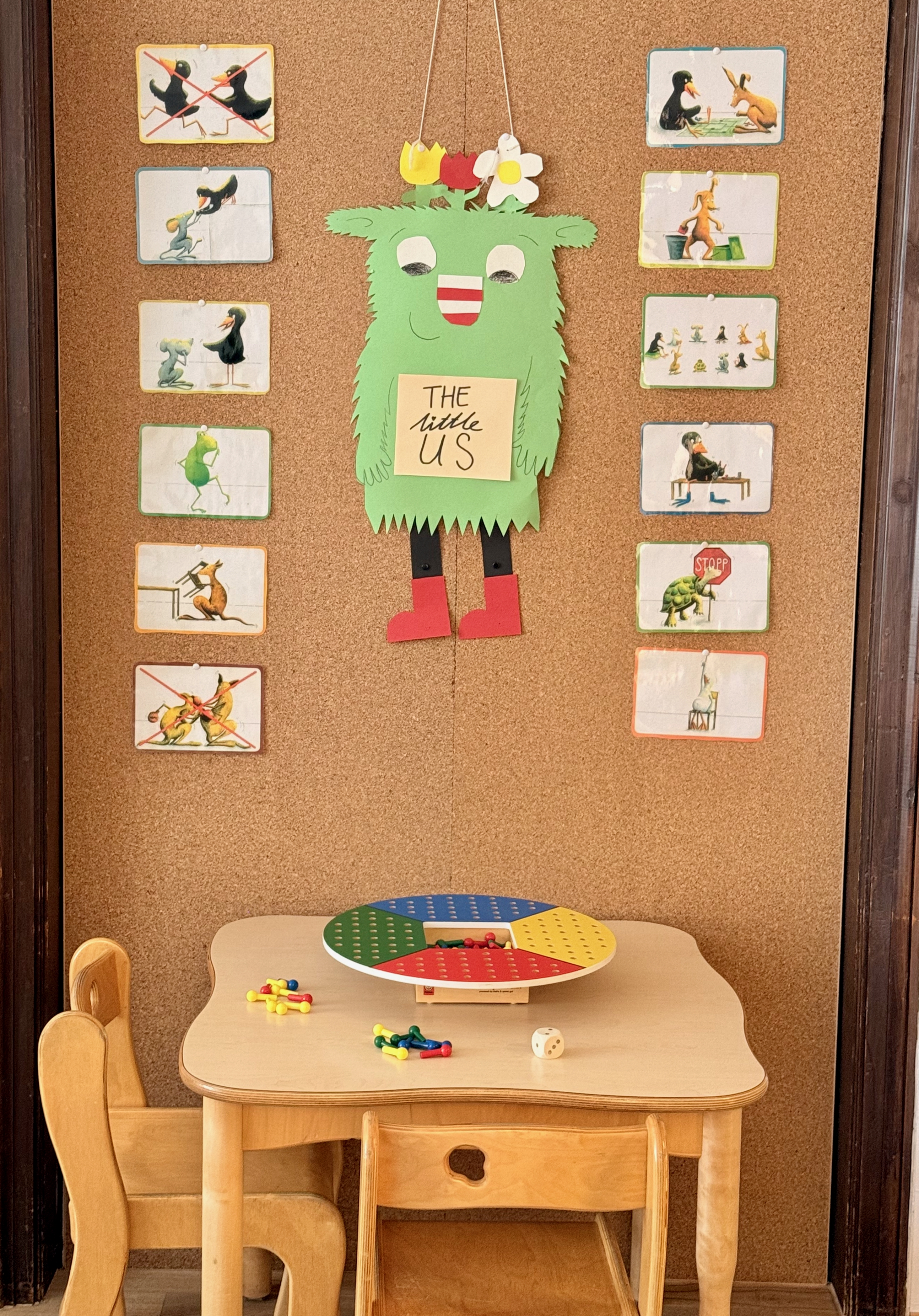 Bastelraum-Ecke mit Pinnwand: ein grünes Monster aus Papier mit der Aufschrift „The little us" in der Mitte, umrahmt von Bildkarten. Davor ein Holztisch mit einem bunten Lernspiel-Kreisel und kleinen Holzstühlen.