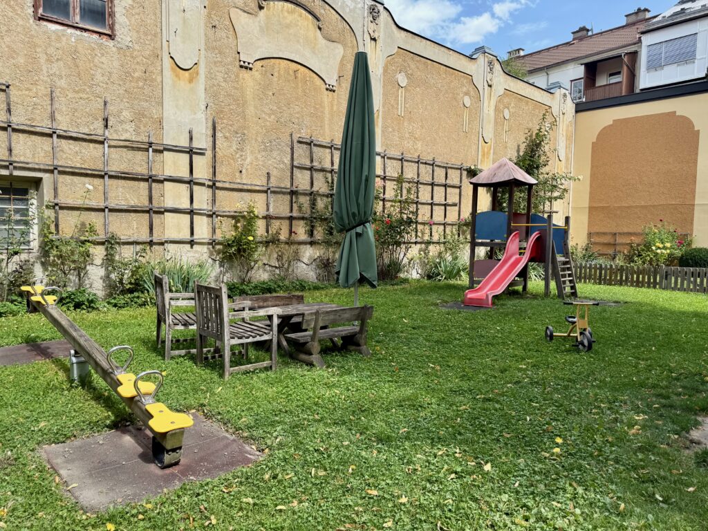 Gepflasterter Garteninnenhof mit gelber Wippe, einer Holzbank-Tisch-Kombination, einer roten Rutsche mit kleinem Klettergerüst und einem Dreirad, umgeben von Grünflächen und einer alten Hausmauer.