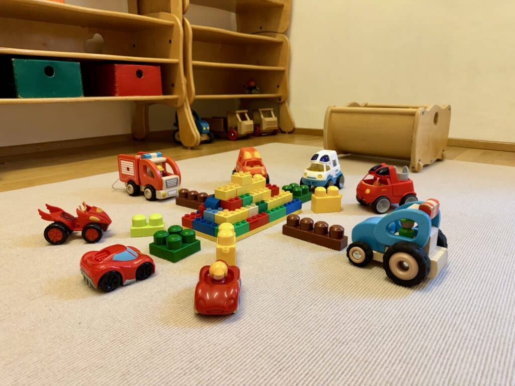 Buntes Spielzeug am Boden ausgebreitet: Bausteine zu einem kleinen Berg gestapelt, mehrere Spielzeugautos, ein Feuerwehrauto und ein Traktor, im Hintergrund niedrige Holzregale.