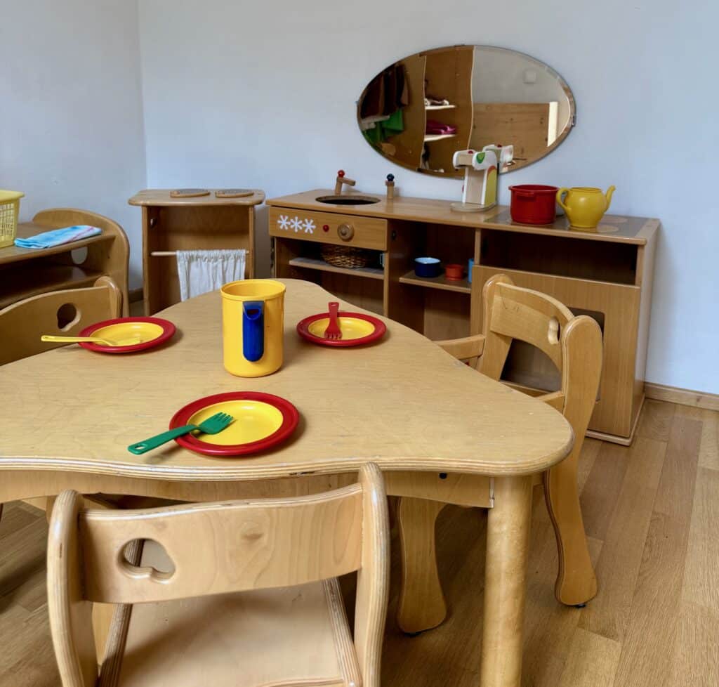 Gedeckter Kindertisch mit gelb-rotem Kindergeschirr (Teller, Becher, Gabel), im Hintergrund eine kleine Kinderküche mit Spiegel, Wasserhahn-Attrappe und Ablagefächern.