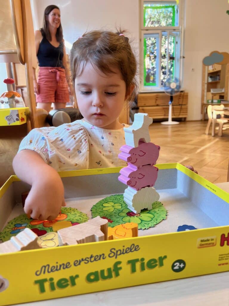 Ein kleines Mädchen spielt konzentriert mit dem Stapelspiel „Tier auf Tier", im Hintergrund steht lächelnd eine Betreuerin.