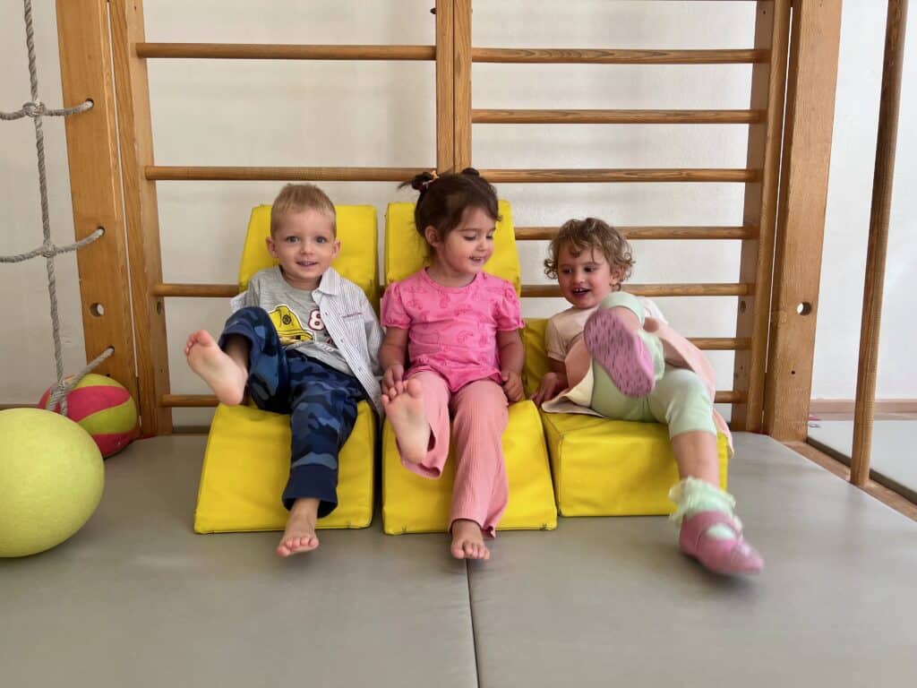 Drei kleine Kinder sitzen lachend nebeneinander auf gelben Sitzwürfeln vor einer Sprossenwand.