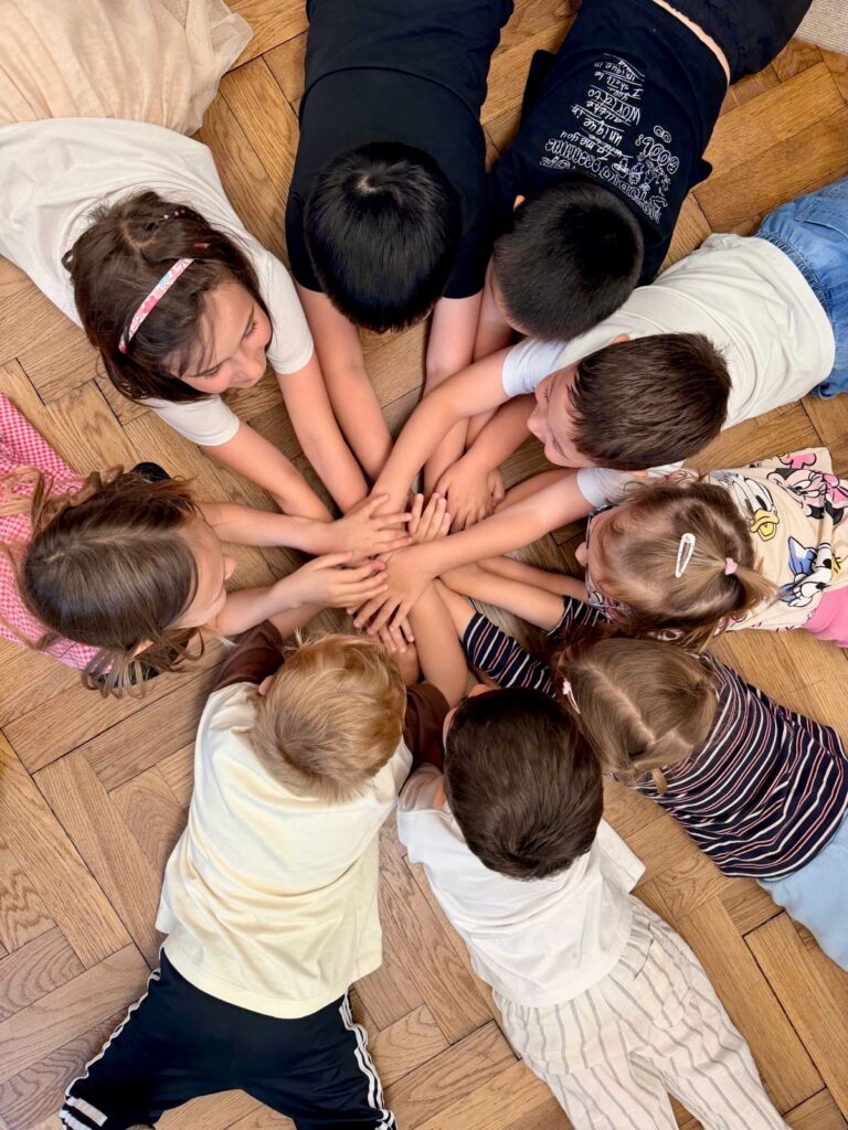 Neun Kinder sitzen im Kreis auf dem Boden und legen ihre Hände in der Mitte zusammen, Aufnahme von oben.