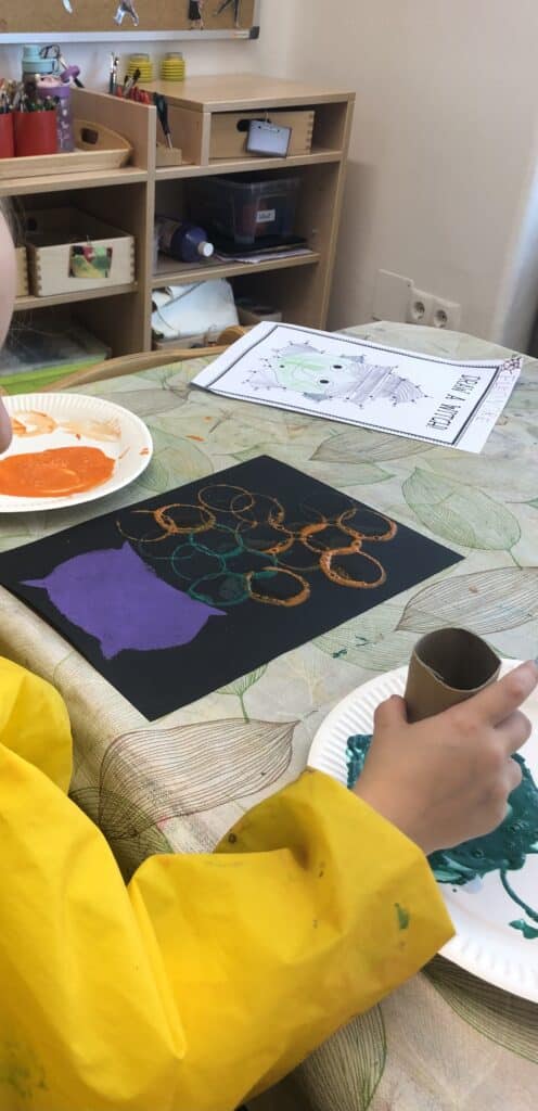 Ein Kind im gelben Malkittel stempelt mit einer Klopapierrolle orange und türkise Kreise auf schwarzes Papier. Sie steigen wie Blasen aus einem lila Hexenkessel auf. Daneben liegen Pappteller mit Farbe und ein ausgemaltes Punkt-zu-Punkt-Blatt mit einer Hexe.