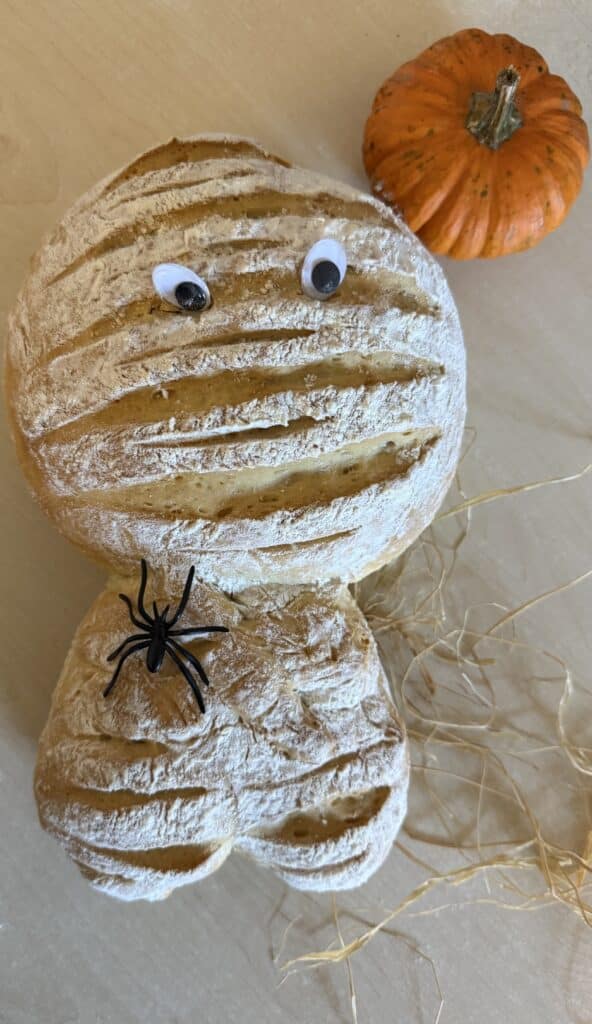 Ein selbst gebackenes Brot in Form einer Mumie. Die Einschnitte in der bemehlten Kruste sehen aus wie Bandagen, dazu kommen Wackelaugen und eine Plastikspinne. Daneben liegen ein kleiner Kürbis und etwas Bast.
